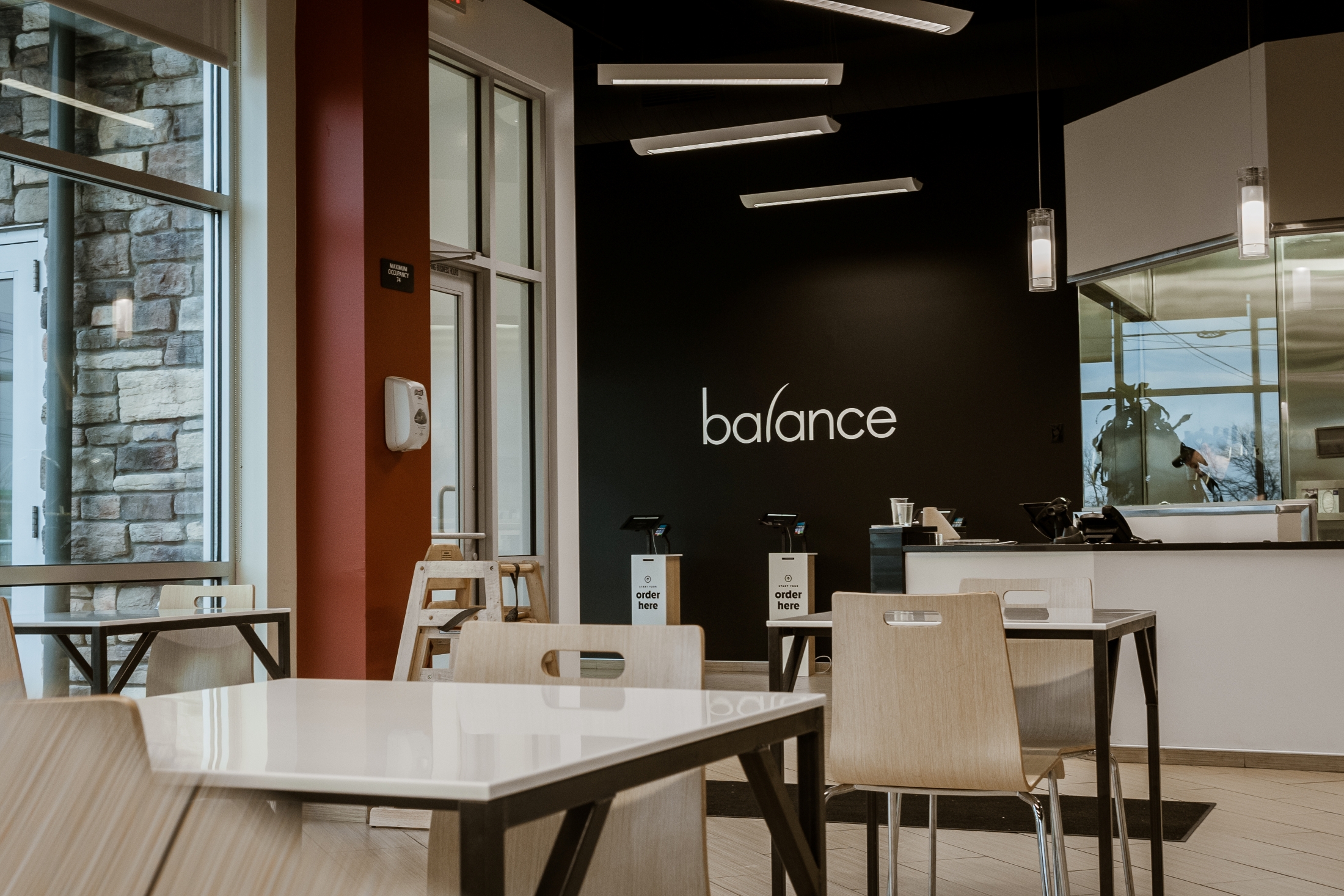 The dining area of Balance Grille with light wooden chairs, white tables, and a black wall featuring the "balance" logo.